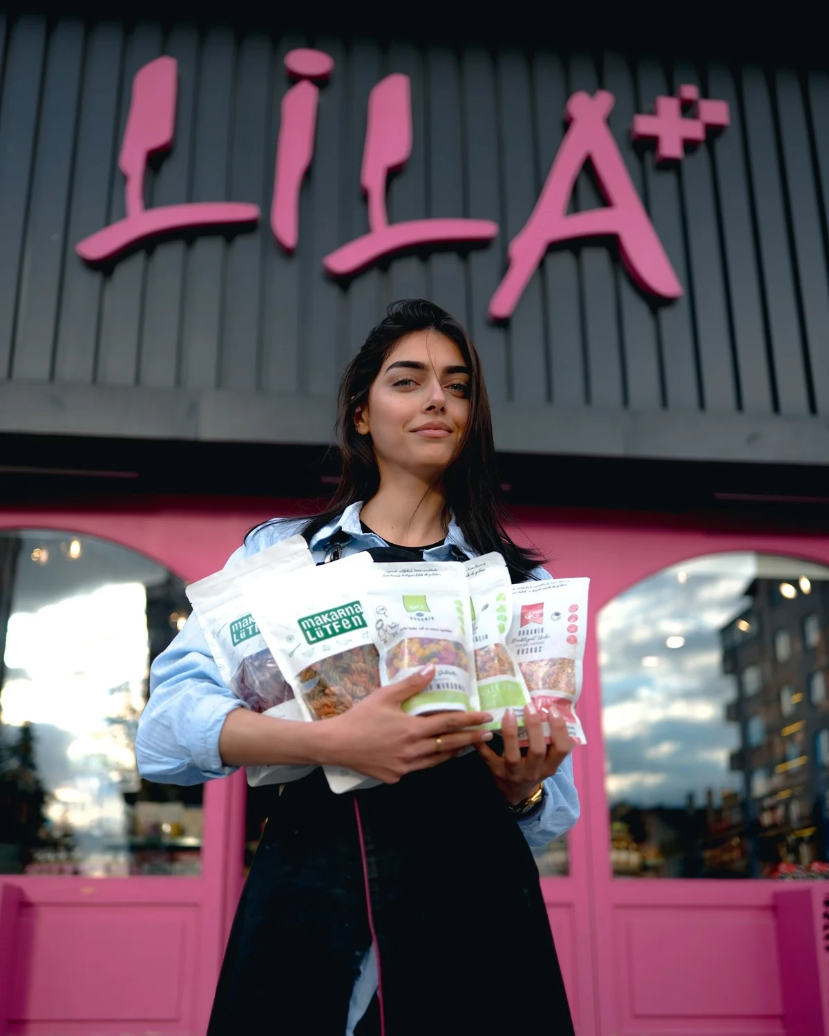 Lila Plus Gastro Market’in pembe tabelası önünde ürünleri taşıyan bir kadın